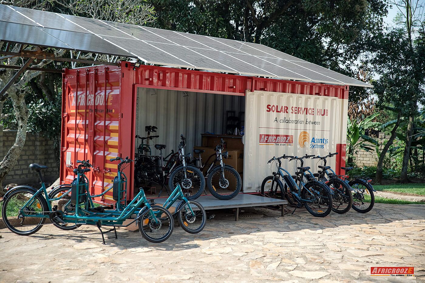 Solar-Ladestation von AfricroozE Ein Schiffscontainer mit der weißen Aufschrift „Solar Service Hub“ steht im Freien unter einem breiten Dach aus Solarpaneelen. Im geöffneten Container und auf dem gepflasterten Platz davor stehen mehrere schwarze und türkisgrüne Elektrofahrräder sowie Lastenräder.