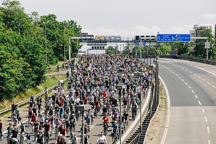 Sternfahrt Berlin 2025 Eine große Menschenmenge fährt auf Fahrrädern über eine mehrspurige Autobahn, die bis zum Horizont mit Radfahrenden gefüllt ist. Über der Straße befinden sich Verkehrsschilder, die unter anderem Richtung „Dresden“ und „Neukölln“ zeigen. Links säumen Bäume die Straße, rechts ist eine leere Gegenfahrbahn zu sehen.